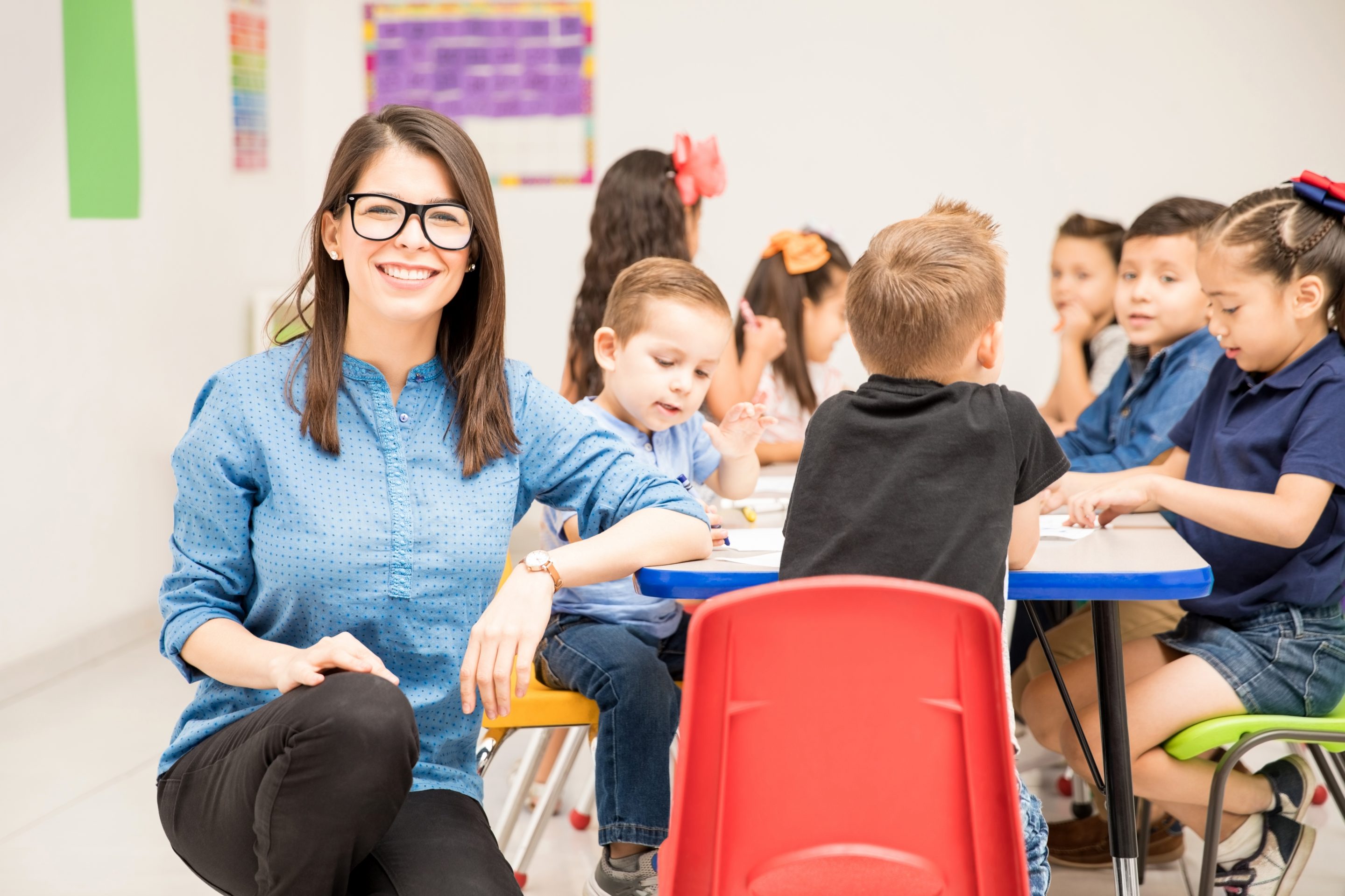 Prevención de riesgos en centros educativos: en la imagen vemos una mujer, profesora de infantil, agachada con el brazo apoyado en una mesa de clase. Lleva gafas y está mirando hacia la cámara y sonriendo. En la mesa hay niños sentados dibujando.