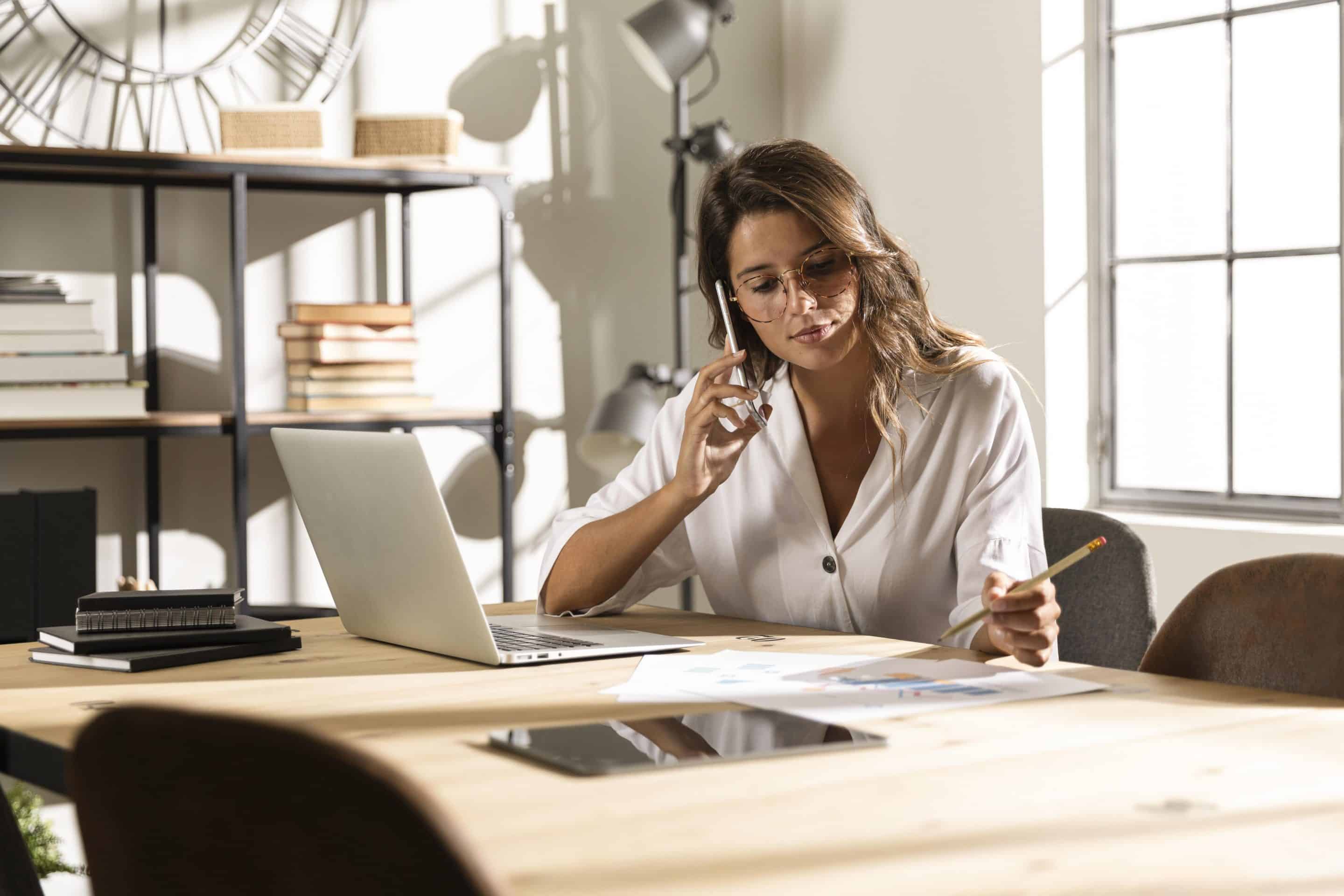 Mujer trabaja sentada delante de la mesa de ordenador en su casa. Lleva gafas y está hablando por teléfono mientras anota algo en una libreta. Imagen para ilustrar el artículo sobre los nuevos riesgos de las plataformas digitales.