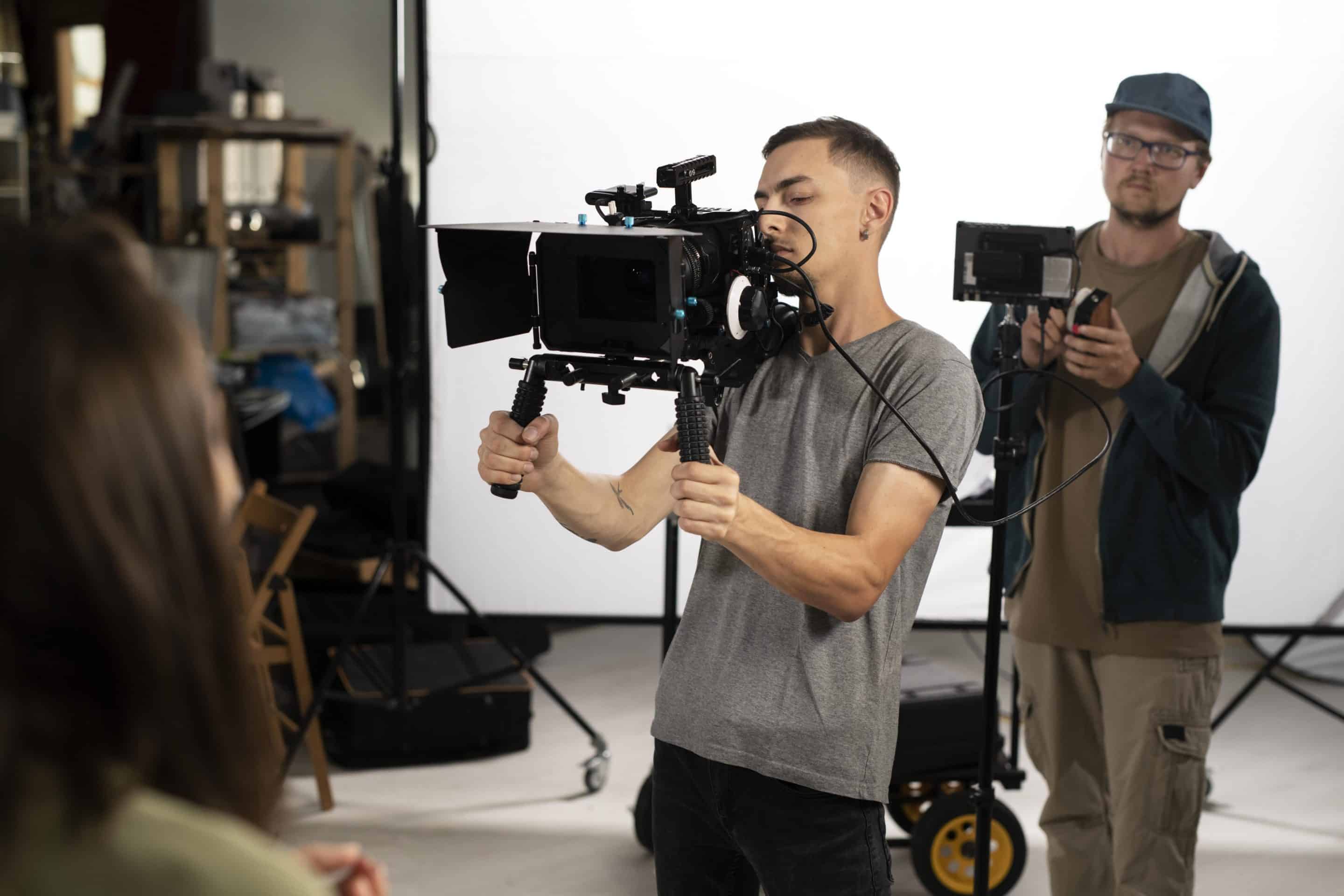 Prevención de riesgos laborales en el sector audiovisual. En la imagen vemos dos hombres jóvenes trabajando en un estudio de televisión, uno de ellos sujerando una cámara de grabación y el otro, detrás, sujetando un foco de luz.