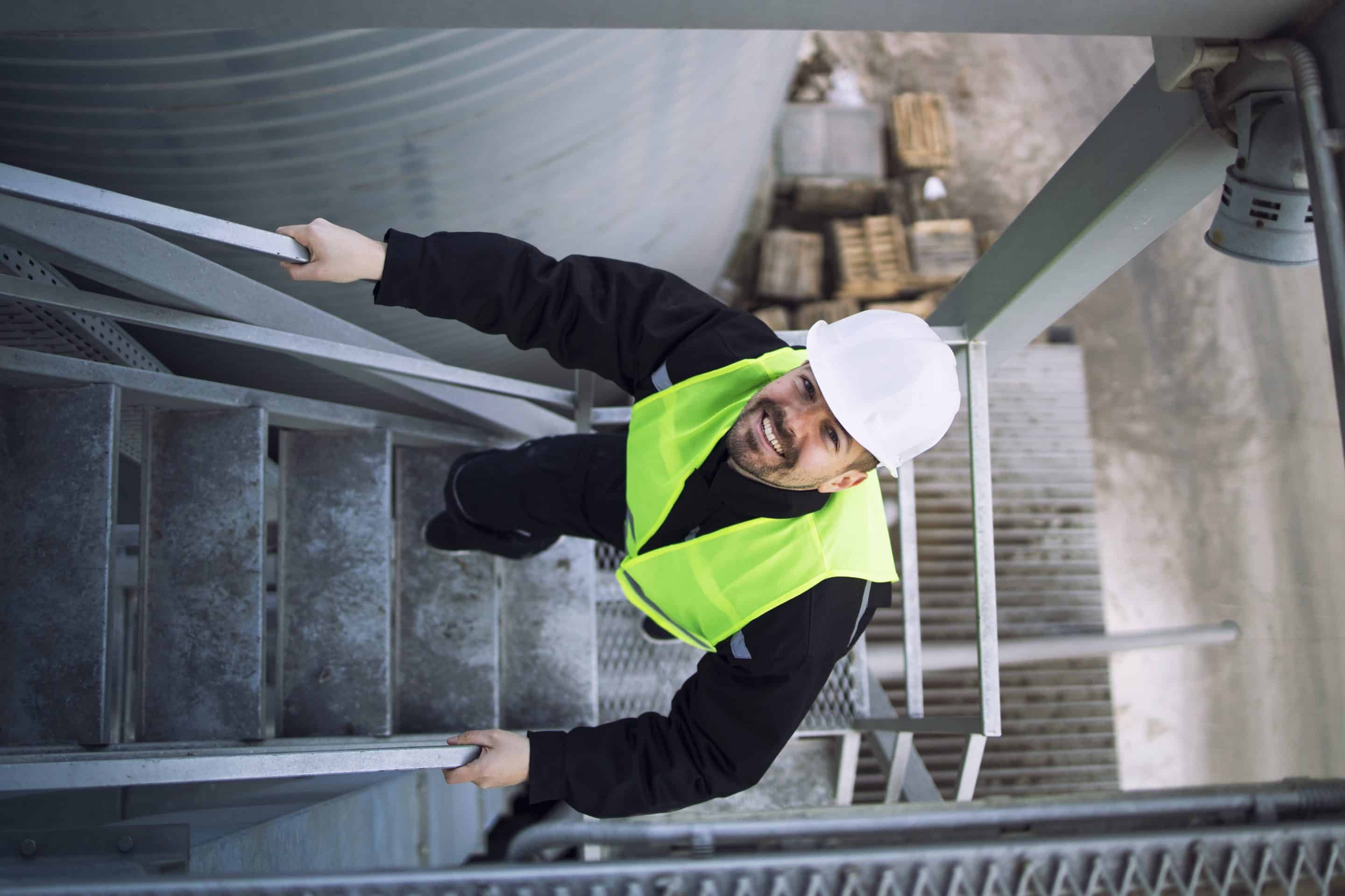 Top view of factory worker climbing metal stairs on industrial silo building. Un trabajador subiendo una escalera parece agobiado por los EPIS y el gesto de su rostro muestra falta de visibilidad debido al sol