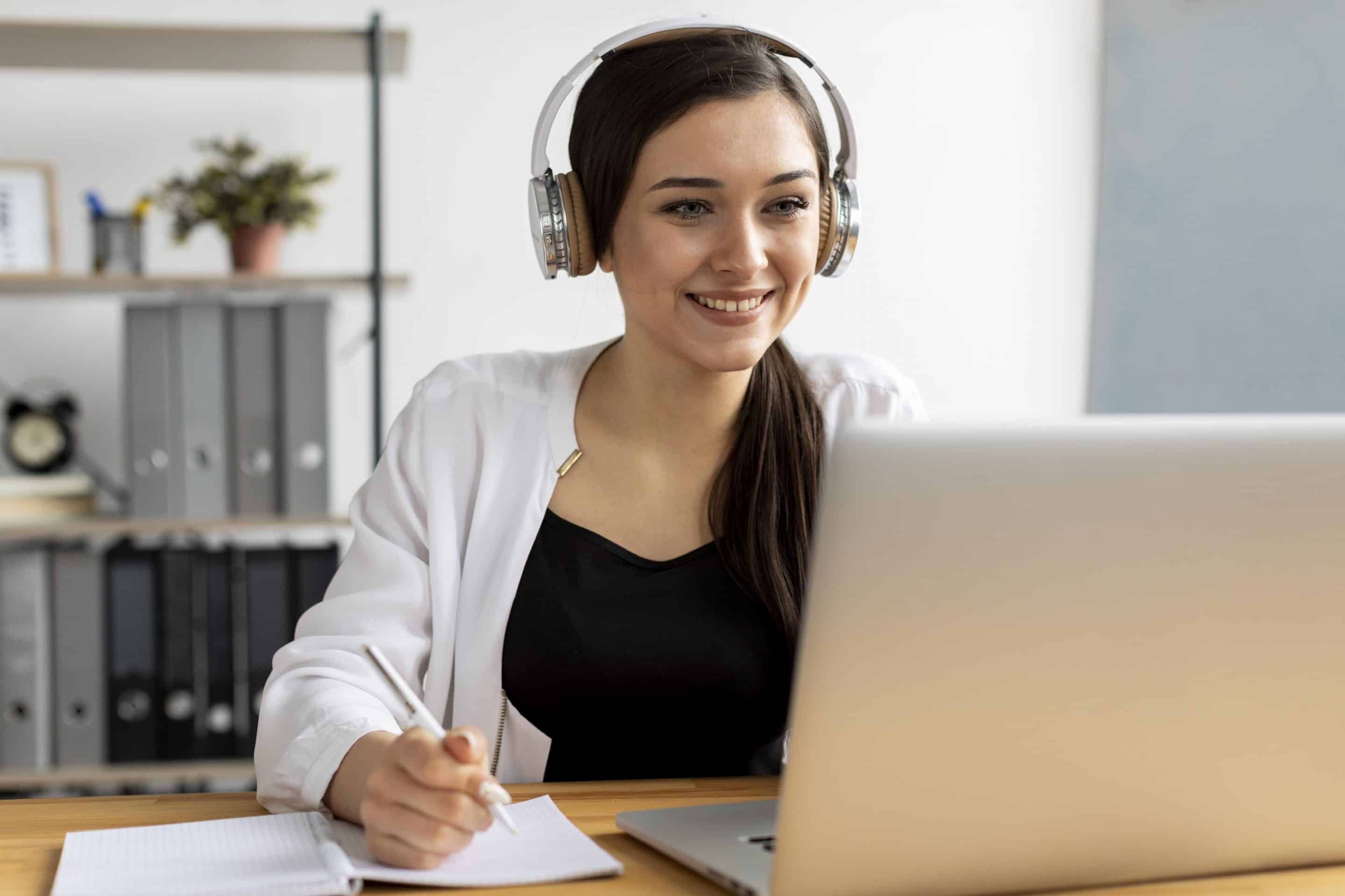 Una mujer sentada al escritorio frente a su portátil, sonriendo y con cascos, tomando notas.