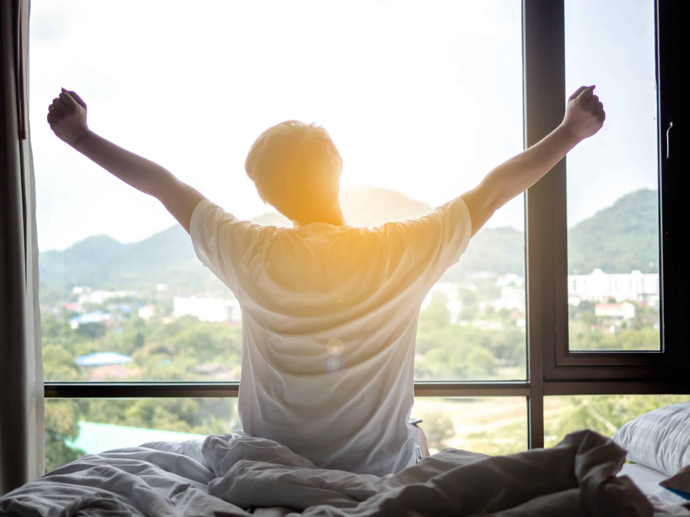 A person waking up and stretching in soft morning light, sitting on the edge of the bed or standing by a window. The scene is calm and quiet, showing the first moment of the day. Natural lighting, no visible technology, a subtle sense of body awareness or tension.