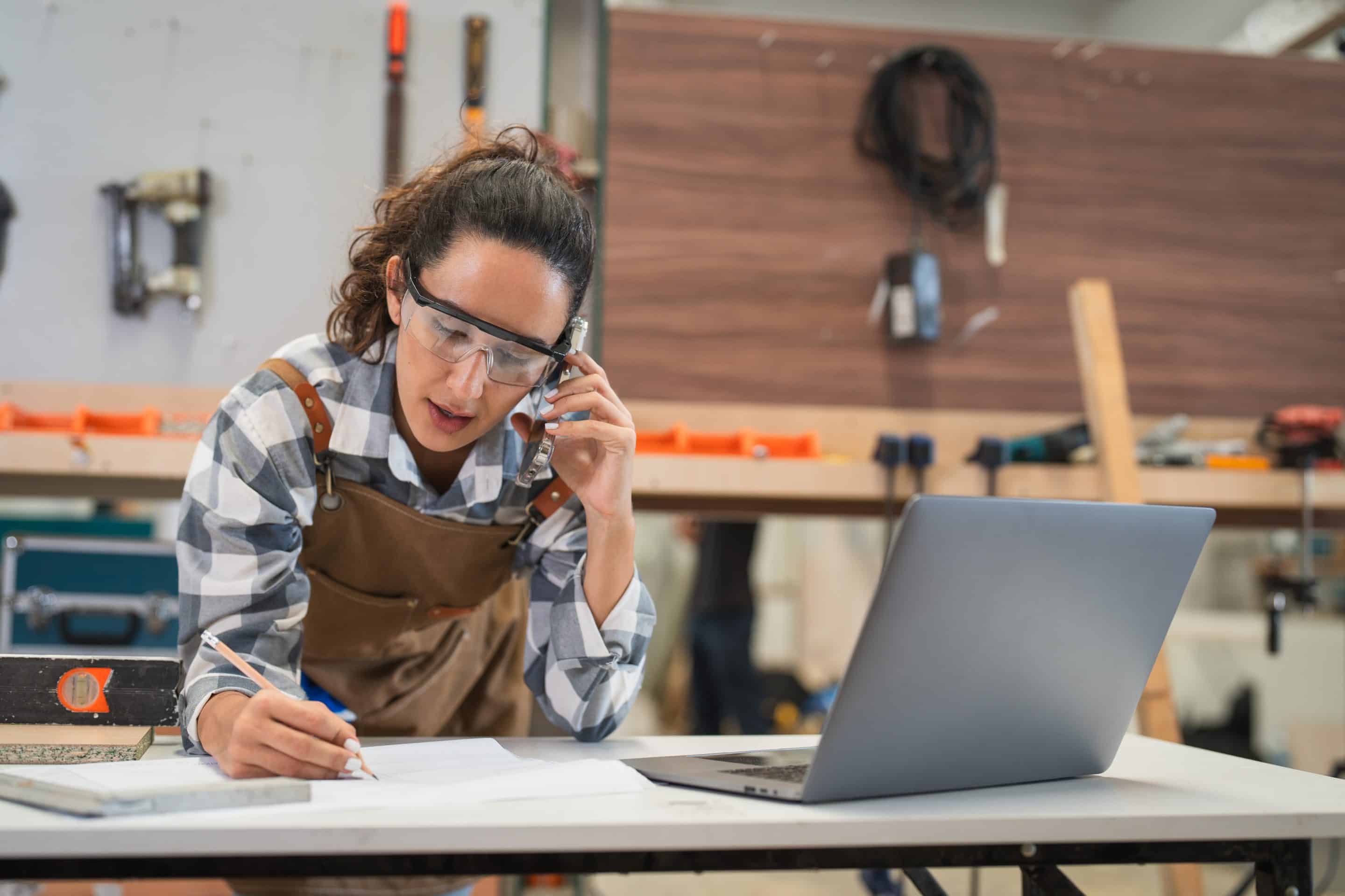 Mujer usando un portátil y hablando por teléfono con un cliente en un taller de carpintería. LLeva gafas y ropa de protección. Muestra la importancia de reforzar la prl en las pymes.
