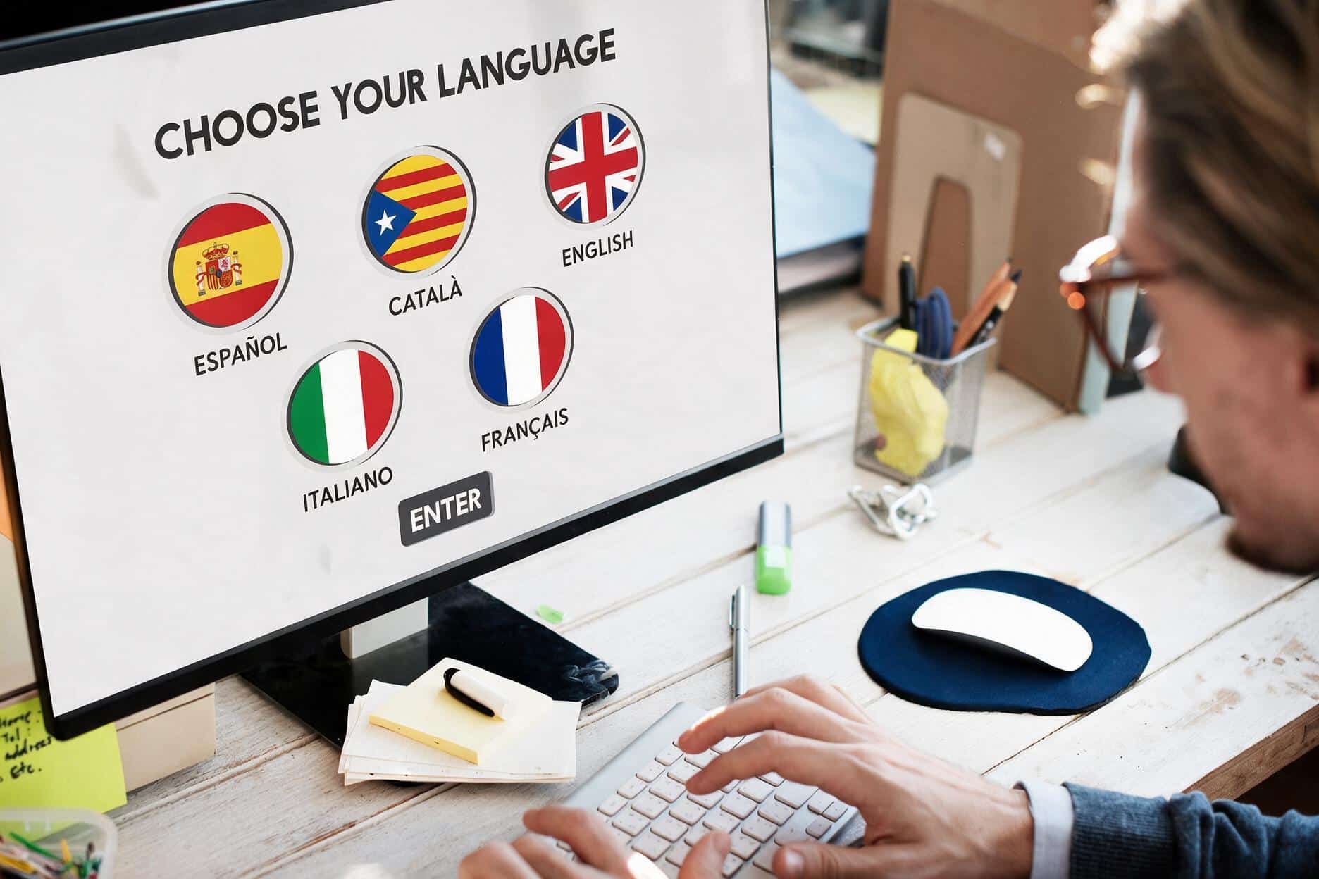 Una persona está sentada frente a una pantalla de ordenador, en la que se muestra un menú que permite seleccionar el idioma, entre inglés, francés, italiano, español o catalán. La imagen muestra la utilidad de una plataforma de formación prl en varios idiomas