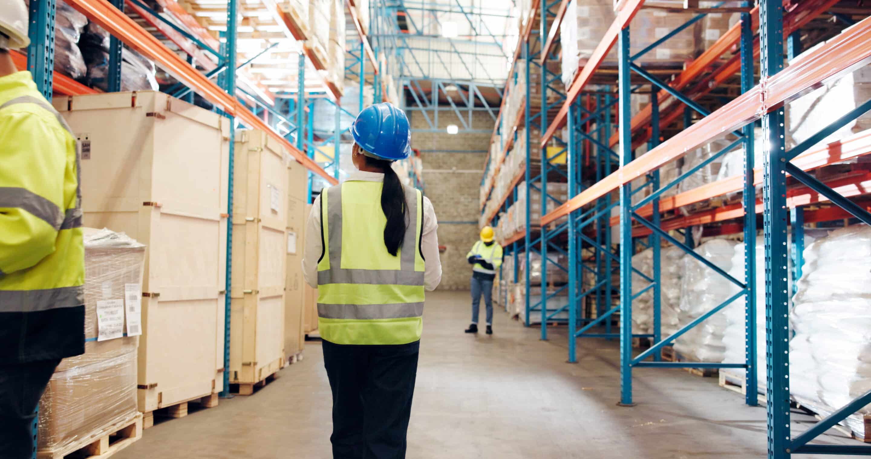 Trabajadores con casco y chaleco de seguridad en un almacén logístico con estanterías y mercancías.